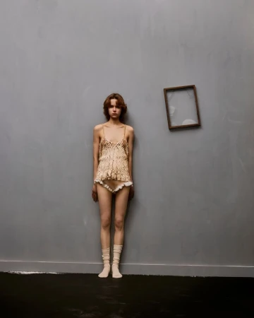 Young woman in lacy lingerie stands against a gray wall, near a floating frame, in a minimalist fashion shot with soft light.