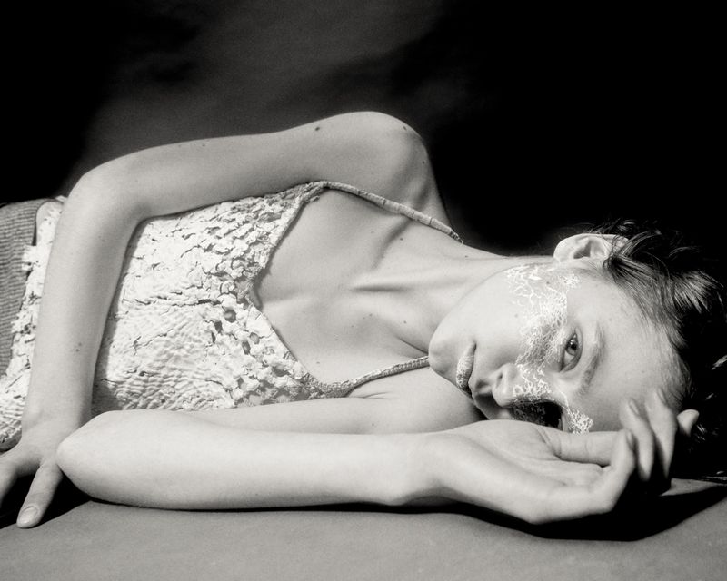 Monochrome portrait of a fair-skinned young adult woman with face paint lying on a flat surface against a dark background.