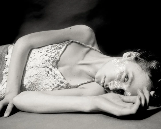 Monochrome portrait of a fair-skinned young adult woman with face paint lying on a flat surface against a dark background.