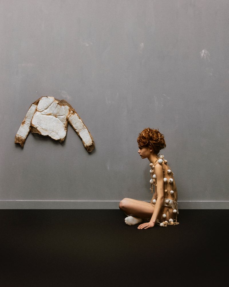 Woman seated indoors wearing beaded garment next to a jacket hanging on the wall, creative fashion, studio lighting and grey background.