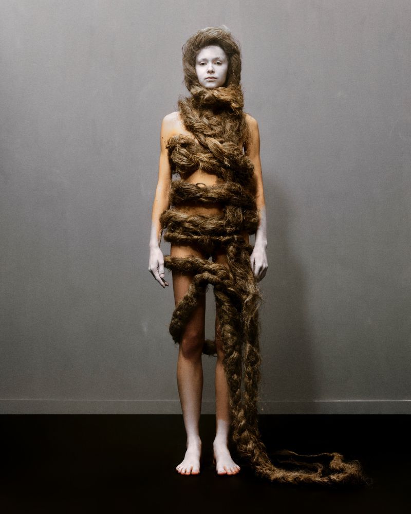 Artistic studio portrait of a young adult woman covered in a long, thick brown braid against a grey background with black floor.