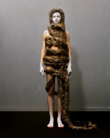 Artistic studio portrait of a young adult woman covered in a long, thick brown braid against a grey background with black floor.