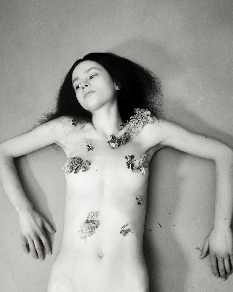 Striking monochrome image features a female nude adorned with floral elements, lying against a muted backdrop in a fine art aesthetic.