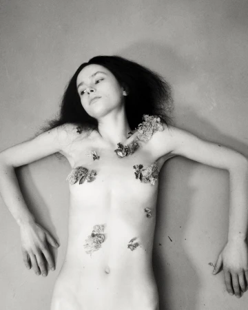 Striking monochrome image features a female nude adorned with floral elements, lying against a muted backdrop in a fine art aesthetic.