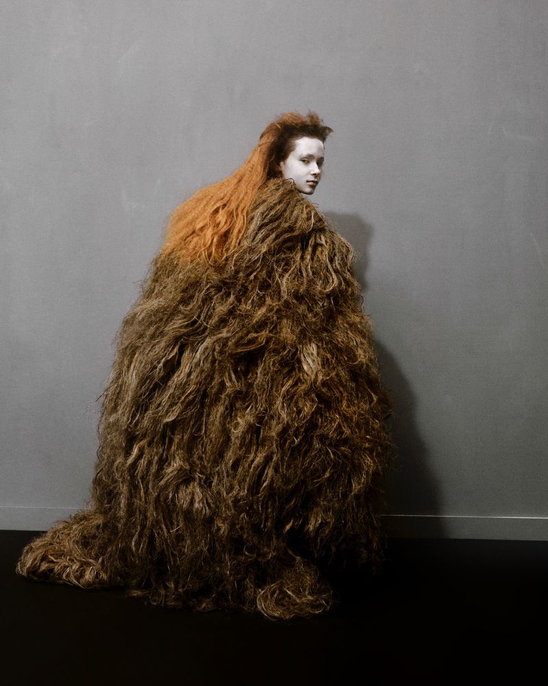 Person in extravagant hair-like costume with reddish-brown shades, looking over shoulder in front of grey wall and dark floor.