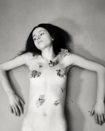 Monochrome portrait of a woman lying down with moss and natural elements placed on her body against a textured background
