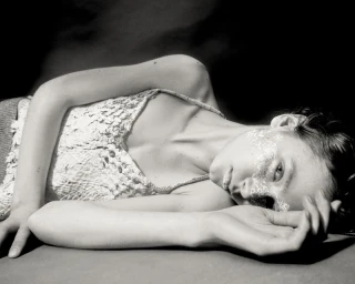Model lying down wearing a textured lace top with intricate white lace makeup on her face against a dark background