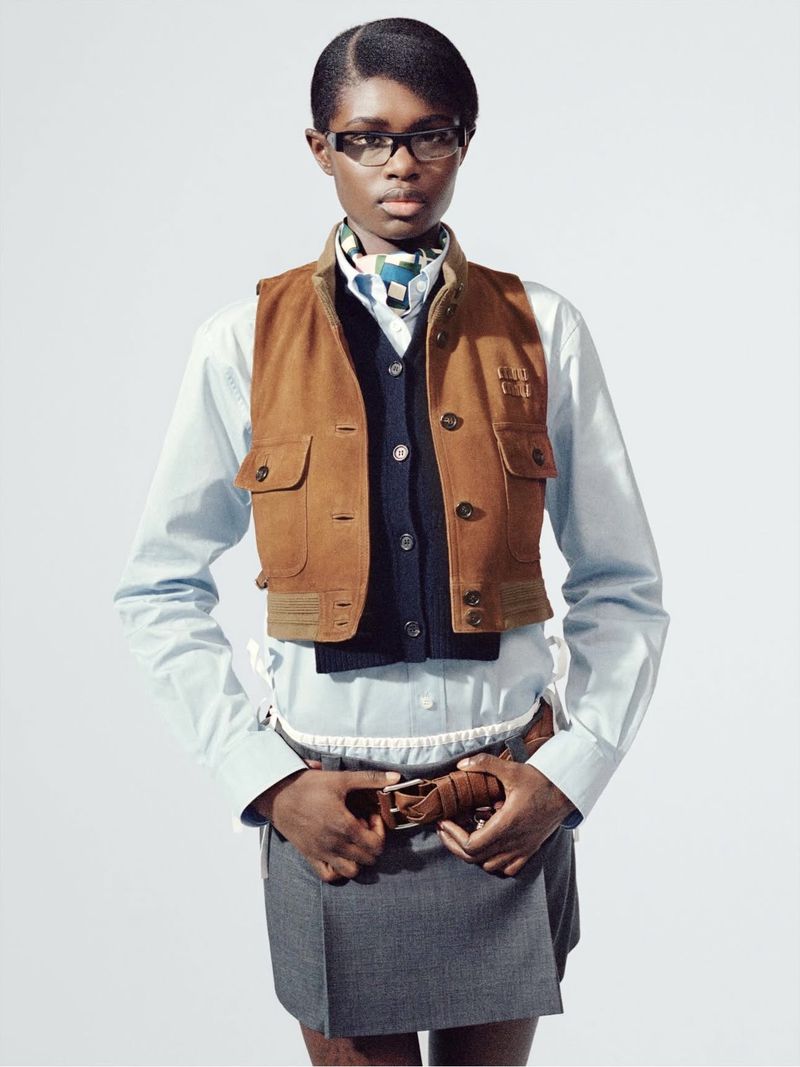 A person wearing a brown vest, light blue shirt, and gray skirt stands against a white background.