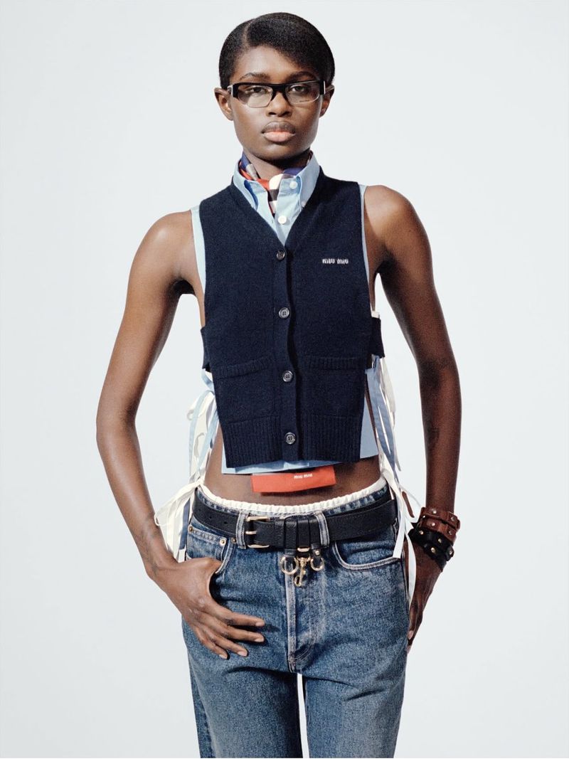 An adult woman poses in a navy vest, blue jeans, a light blue shirt, and glasses in a brightly lit studio for a fashion shot.