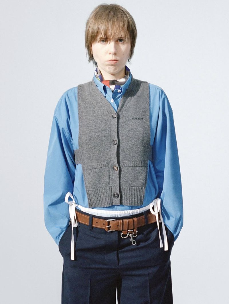 Adult in gray vest, light blue shirt, and navy slacks with brown belt in a studio shot. Full shot, portrait layout, light background.