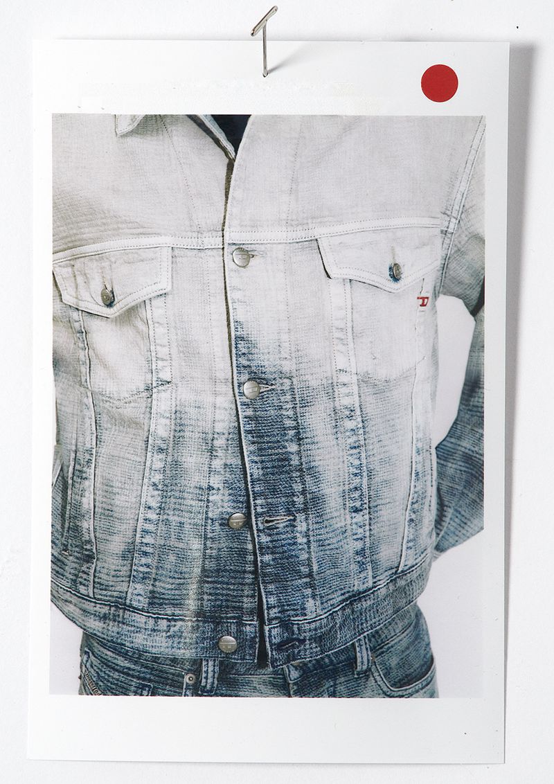 Close-up photo of light-wash denim jacket and jeans attached to a white wall with a metal pin, featuring a red circular sticker.
