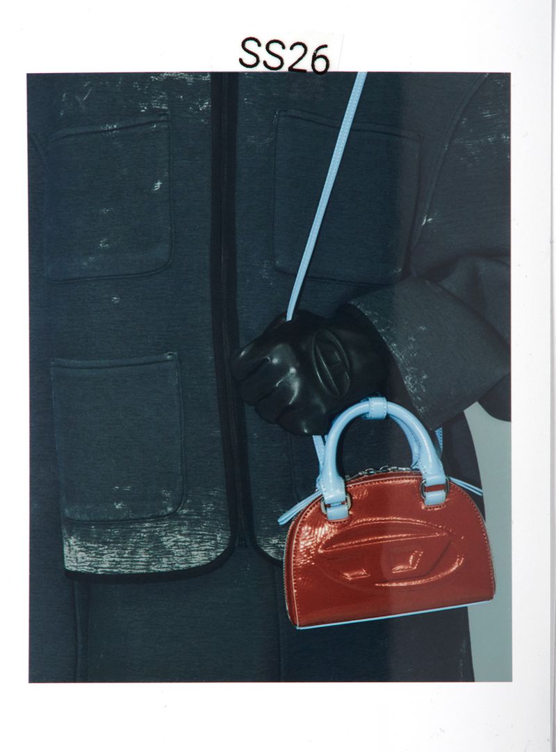 Close-up of a glossy red handbag with light blue handles held against a dark distressed jacket for Diesel SS26.