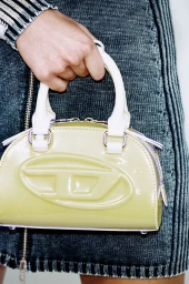 Close-up of a person holding a glossy pale yellow handbag with white top handles and a large D logo against a denim outfit.