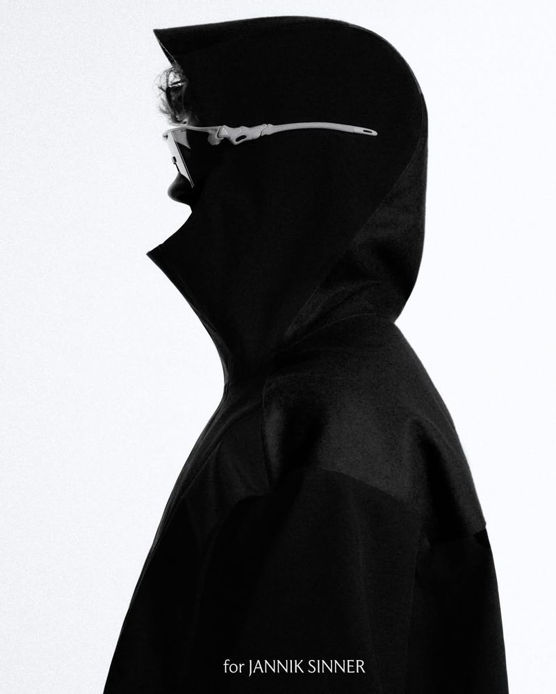 Profile portrait of a person wearing futuristic glasses and a black hooded coat against a bright background, emphasizing fashion and style.