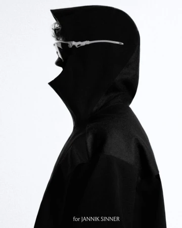 Profile portrait of a person wearing futuristic glasses and a black hooded coat against a bright background, emphasizing fashion and style.