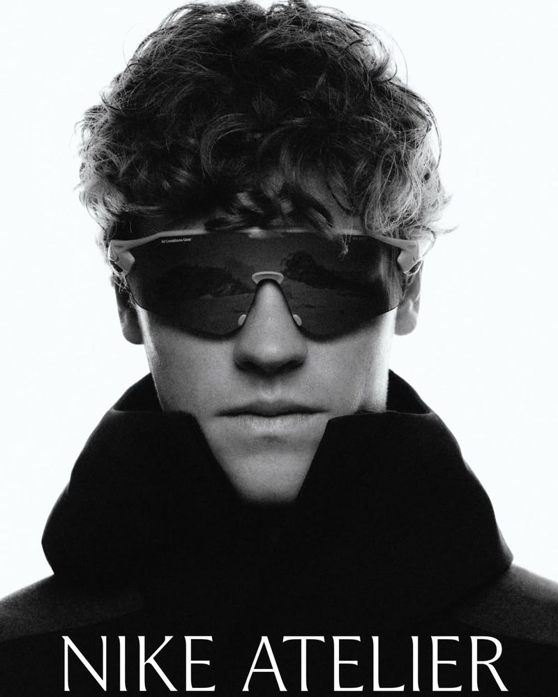 Black and white studio portrait of a curly-haired man wearing sunglasses and a large collar, featuring Nike Atelier branding.