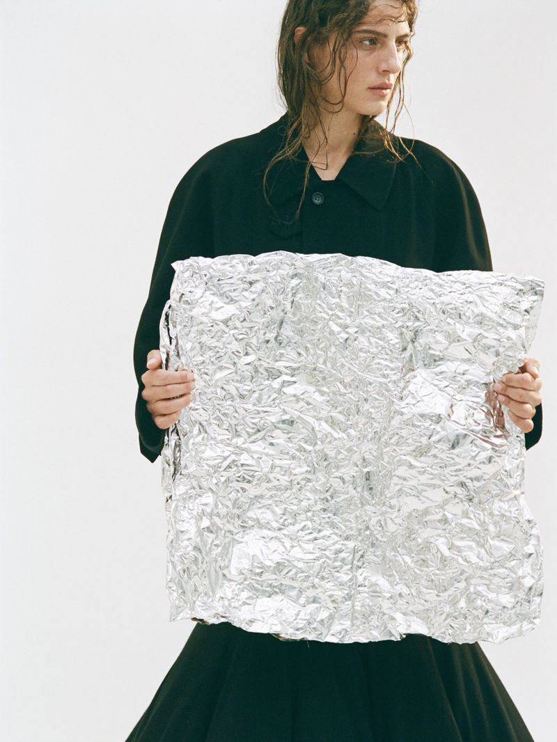 Fashion portrait of a female model holding a large sheet of crinkled aluminum foil in front of a white backdrop with wet hair.