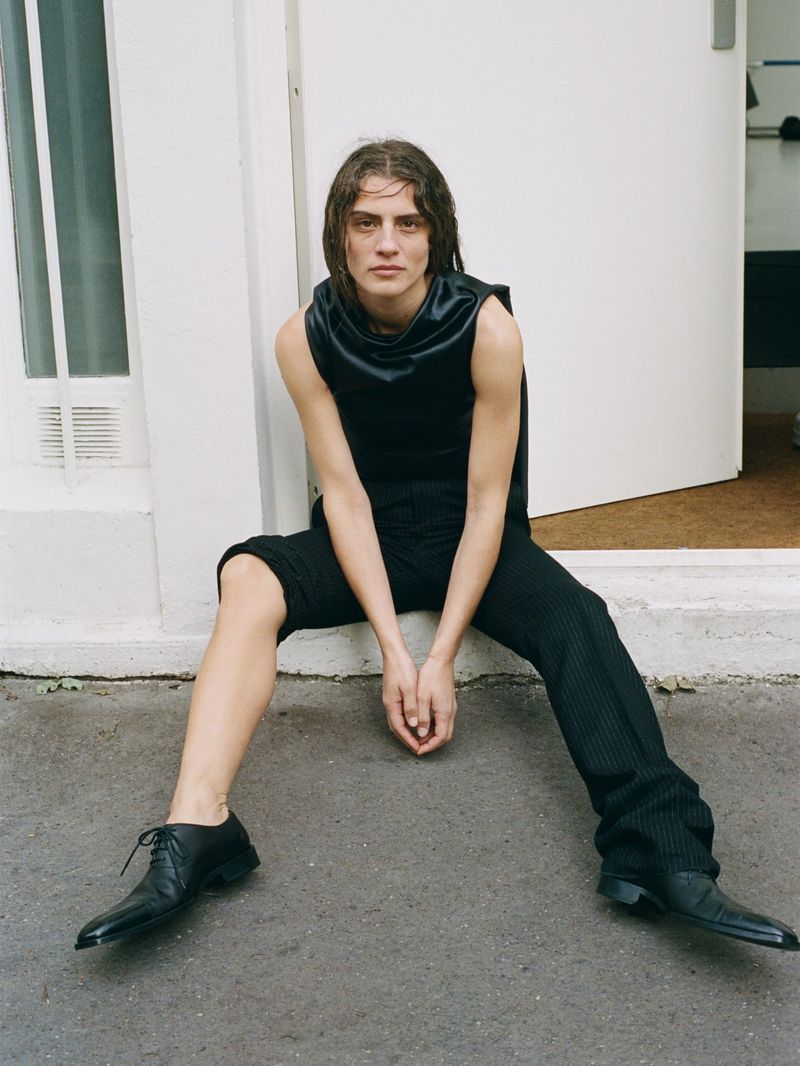 A slender adult wears a black sleeveless top and pinstriped pants, sitting on concrete against a white wall in an outdoor setting.