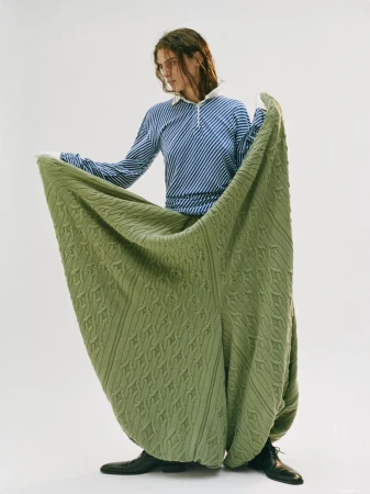 A person in a striped shirt and large green textured pants stands against a white background, showcasing a unique fashion style.