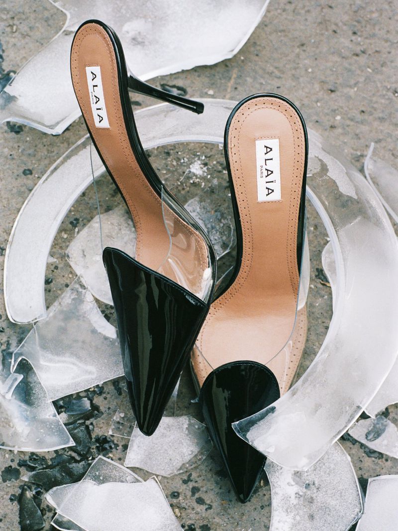 Alaïa high heels in black patent leather and clear plastic, surrounded by shards of broken glass on a grey concrete surface.