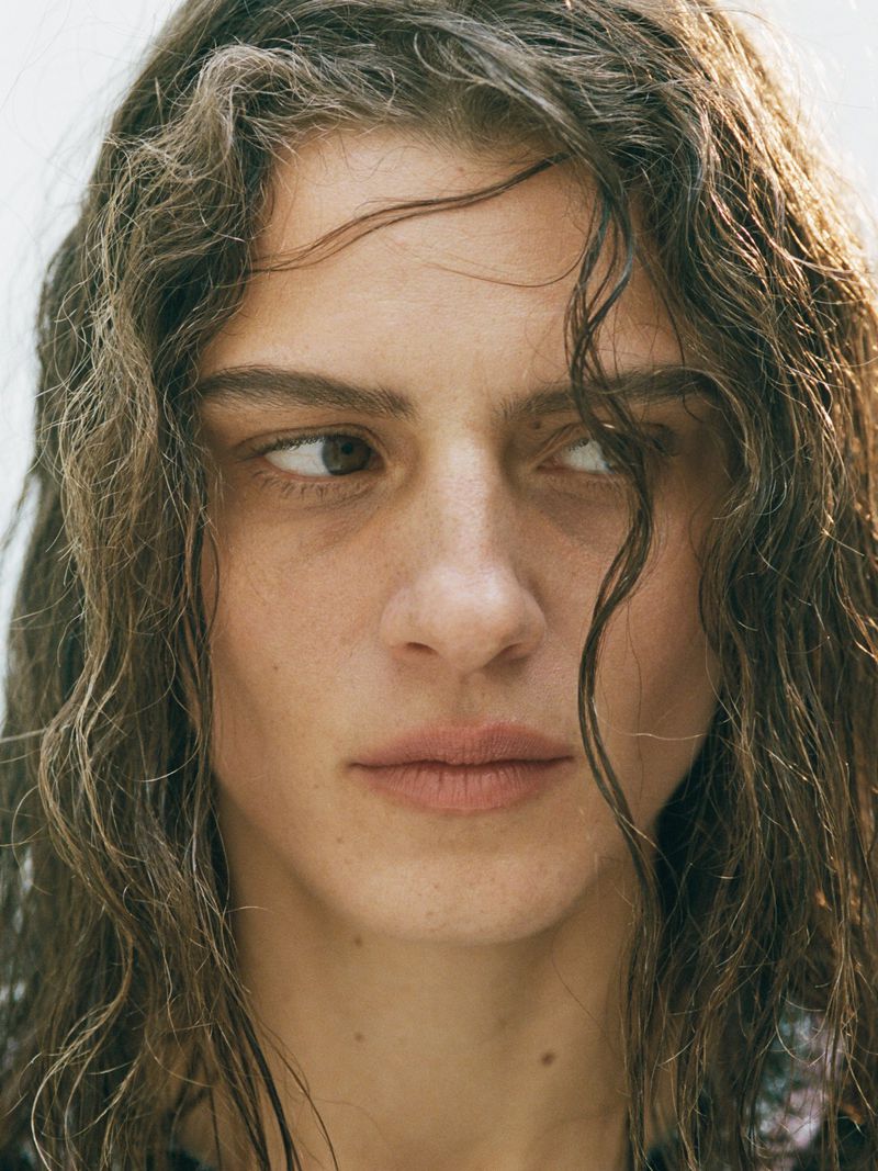 Close-up portrait of a young adult woman with wavy brown hair looking away, with a slightly melancholic expression, against a bright background.