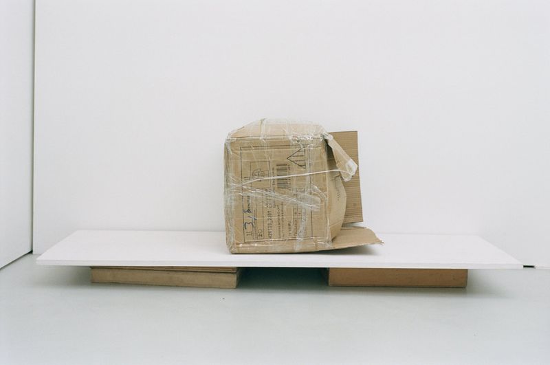 A damaged cardboard box wrapped in clear plastic stands on a white platform against a white wall in a brightly lit indoor setting.