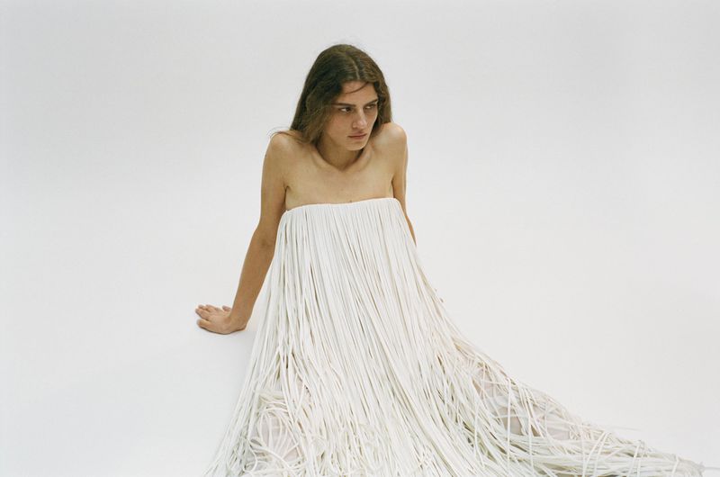 A person wearing a long white dress with fringe details poses in a photo studio with a minimalist, light background.