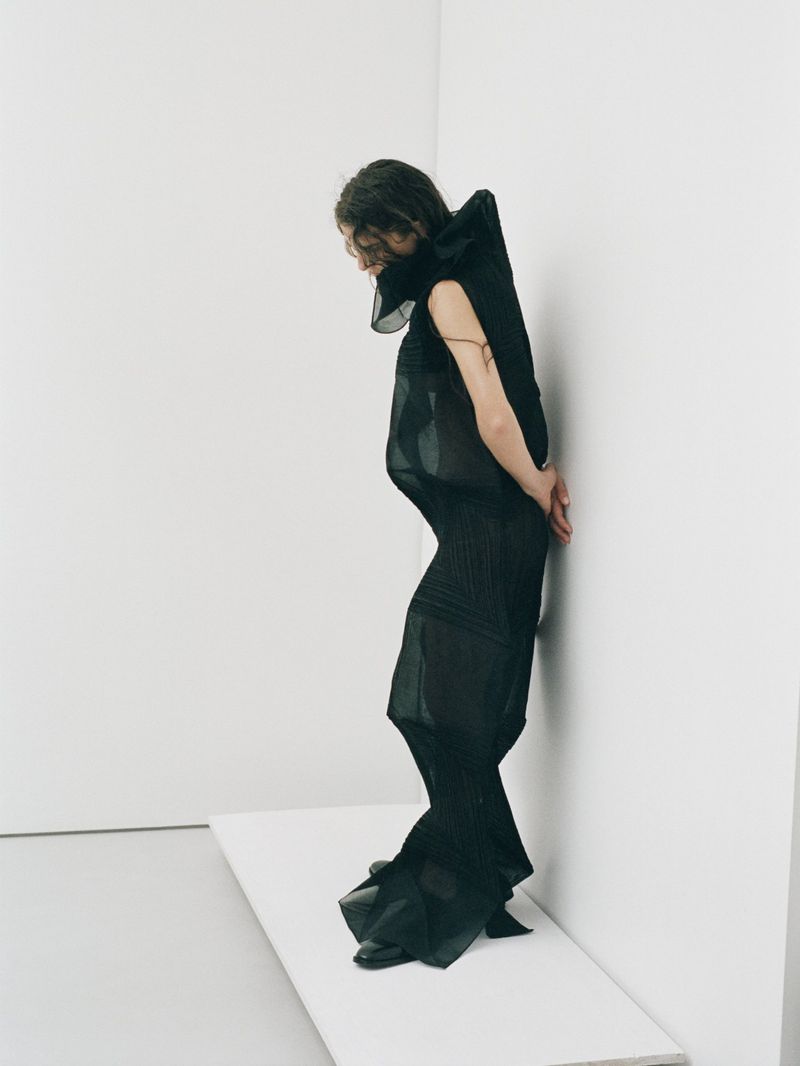 An adult female fashion model in a dark dress stands in a photo studio with a white background looking down with a slight bend at the waist.