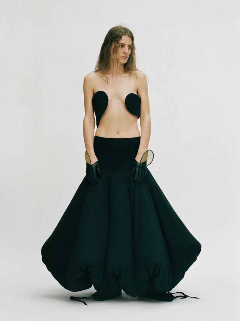 A woman poses in a black top and a voluminous black skirt against a white backdrop, creating a high-fashion and editorial image.
