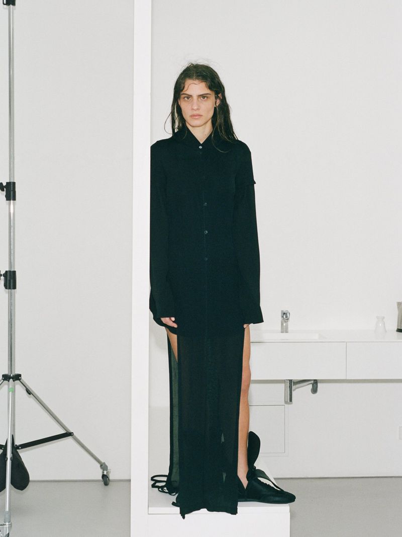 An adult female stands indoors in a black dress, styled for a fashion shoot in a photo studio. White walls and photo equipment visible.