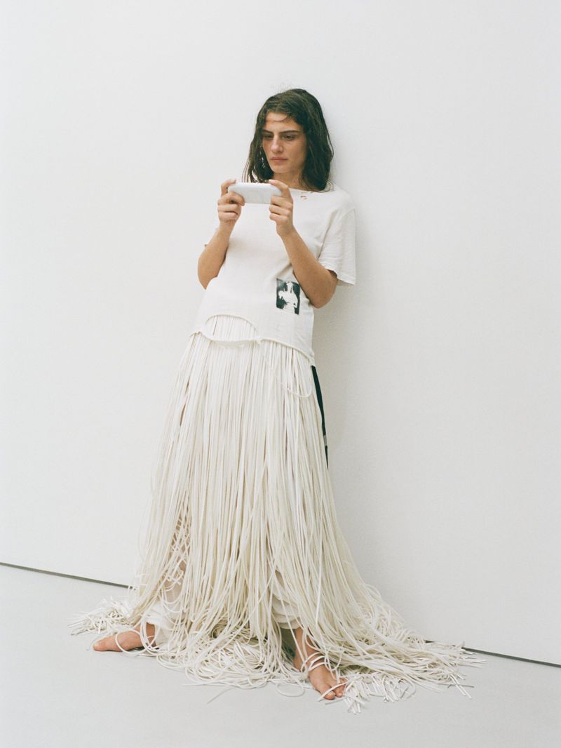 A woman in a white t-shirt and string skirt sits against a white wall while looking at a mobile phone in a well-lit indoor setting.