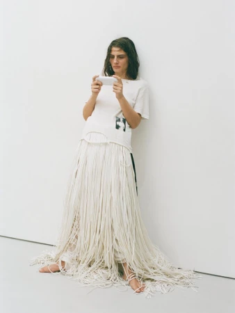 A woman in a white t-shirt and string skirt sits against a white wall while looking at a mobile phone in a well-lit indoor setting.