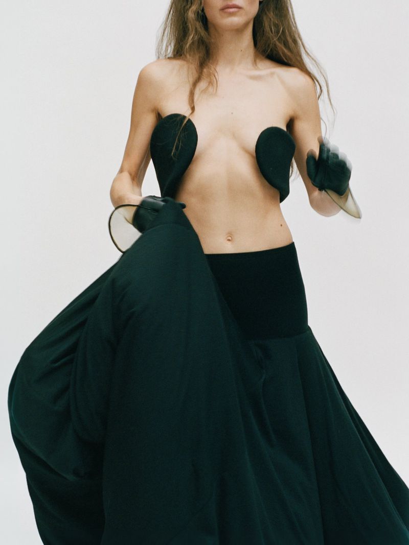 Fashion shot of an adult female model in a dark skirt and sculpted bodice with bare shoulders and black gloves in a white studio.