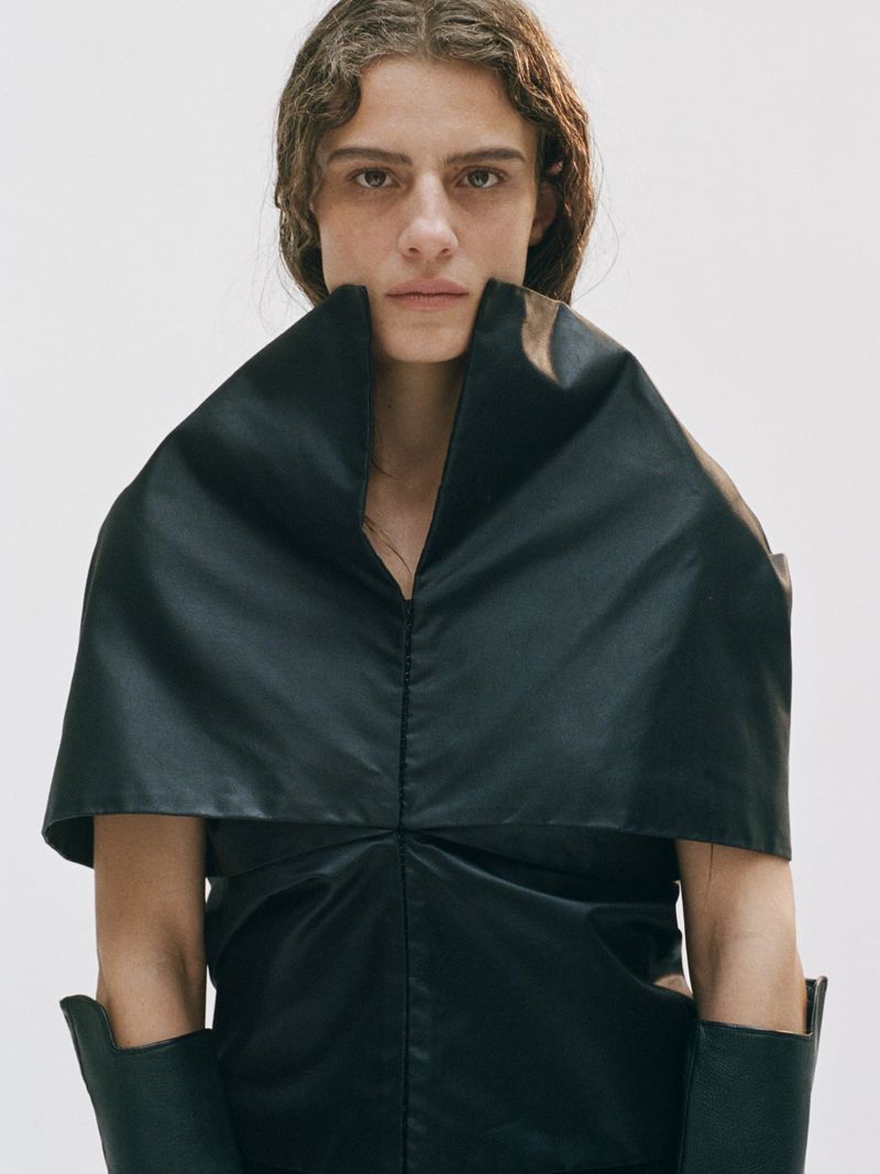 Fashion portrait of a brunette adult female in a black leather dress and matching gloves against a plain white studio backdrop.