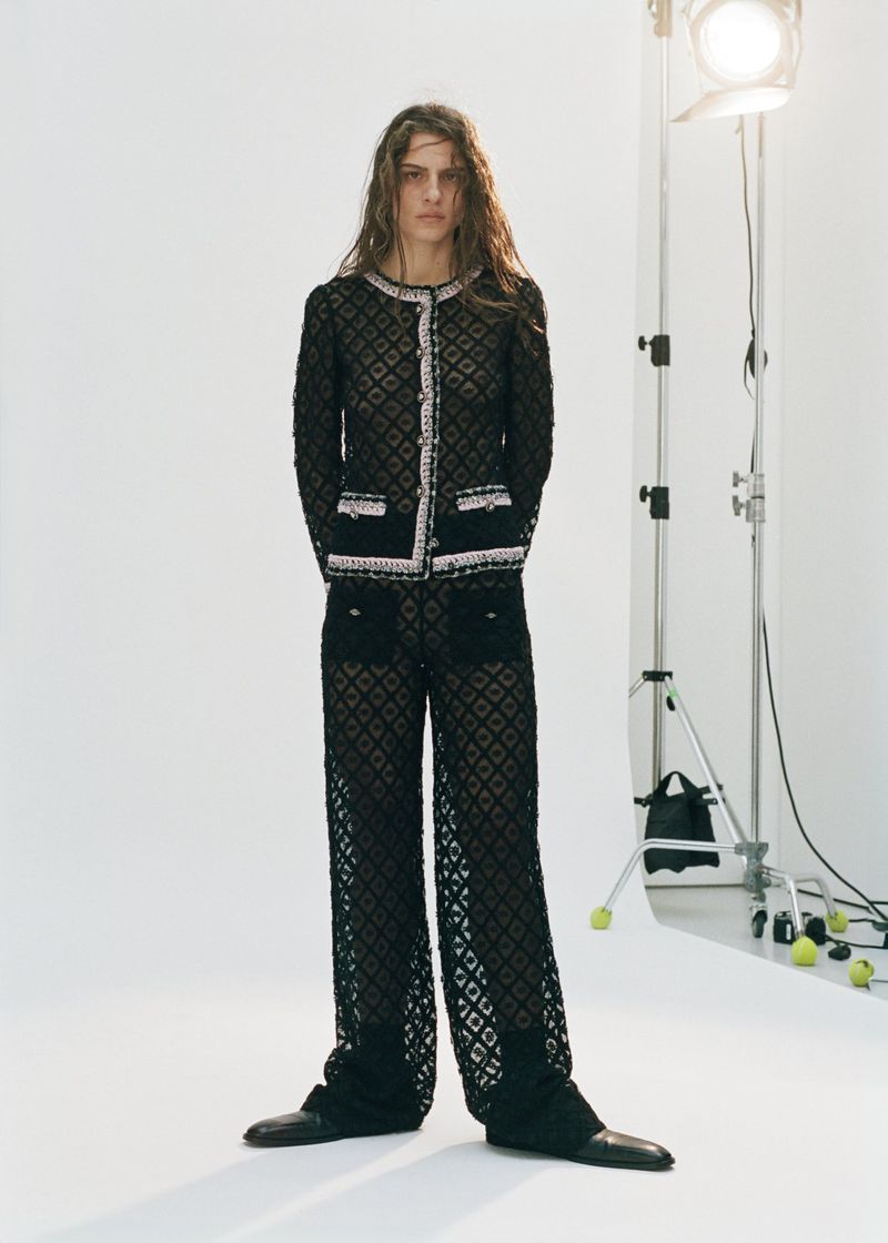 A full shot of a woman standing in a photo studio, wearing a black knit jacket and loose, patterned pants, with studio lighting visible.