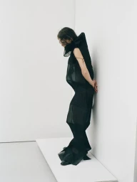 Model in a sculptural black dress standing against a white studio wall for Noisé Magazine