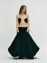 Model wearing a structured black sculptural gown with black gloves against a plain white studio backdrop.
