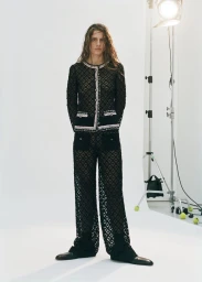 Model wearing a black patterned matching jacket and trousers set standing against a plain white studio backdrop with a spotlight stand.
