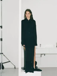 Model standing behind a white studio frame wearing a long black shirt dress against a minimal white interior.