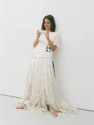 Model wearing a white graphic t-shirt and long fringed skirt holding a white smartphone against a plain wall.