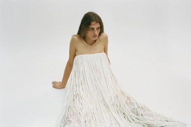 A woman wearing a white fringed strapless dress seated against a plain white studio background.