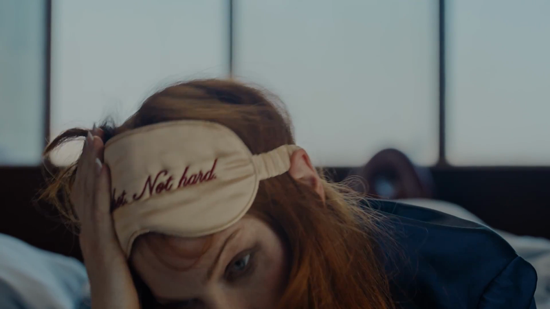 Woman with red hair and eye mask lies in bed in a close-up shot, indoors. Mask reads 'It's Not Hard'.