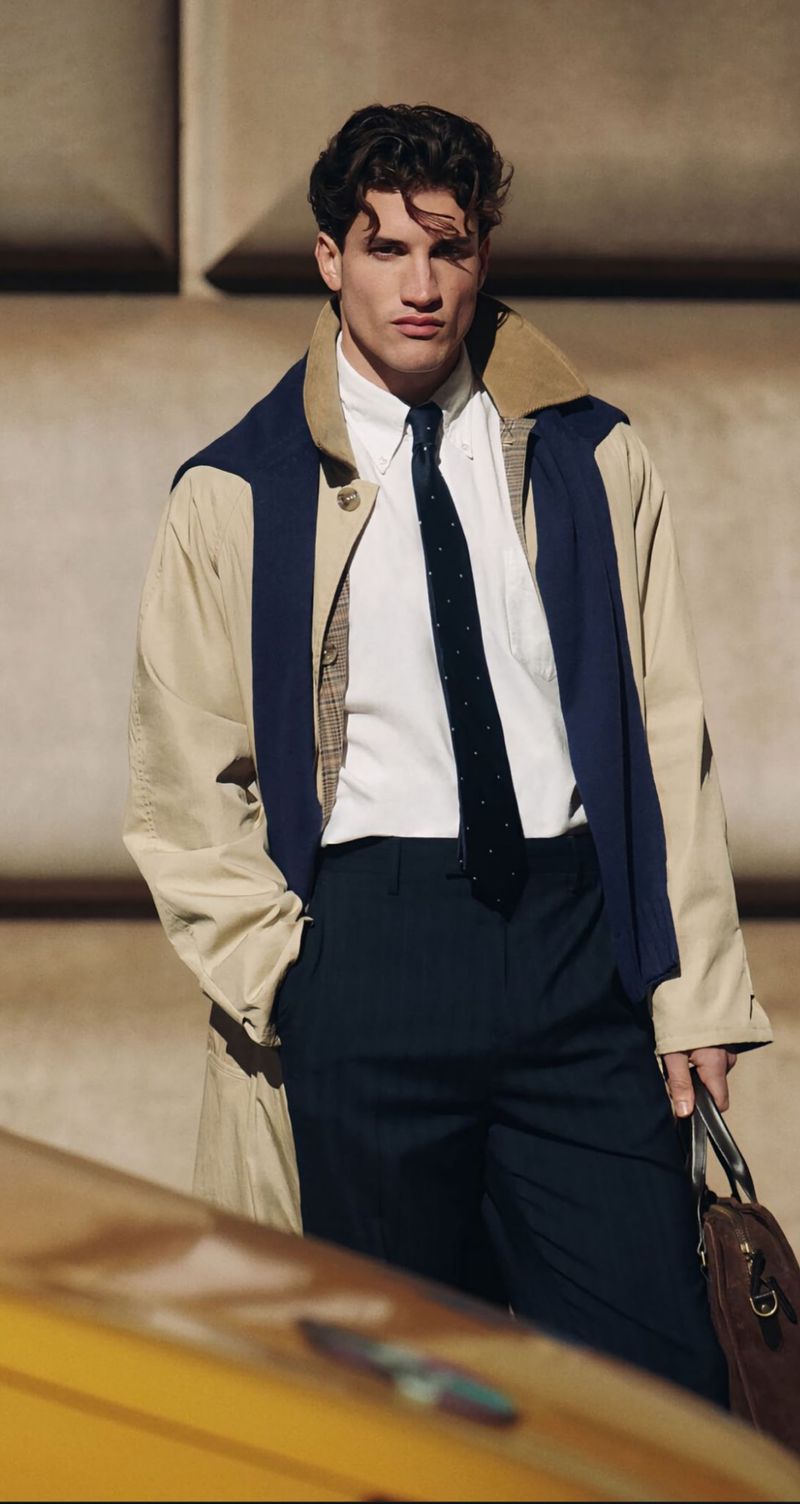 Male model poses wearing tan trench coat, navy suit, white shirt, and navy tie, holding brown leather briefcase against brown background.