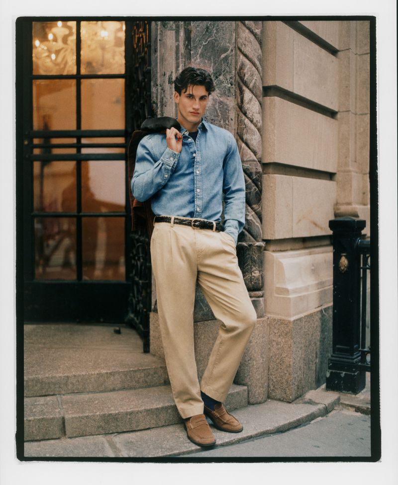 A man in a blue shirt and khaki pants leans against a stone wall next to a door with a brown jacket draped over his shoulder.