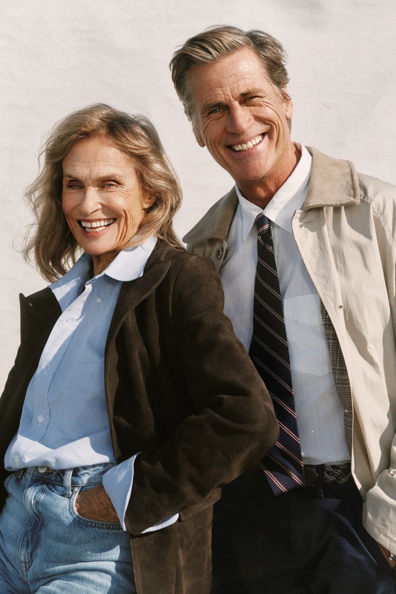 Smiling older couple outdoors: a woman in a brown jacket and jeans, and a man in a beige jacket and tie. Both are happy and relaxed.