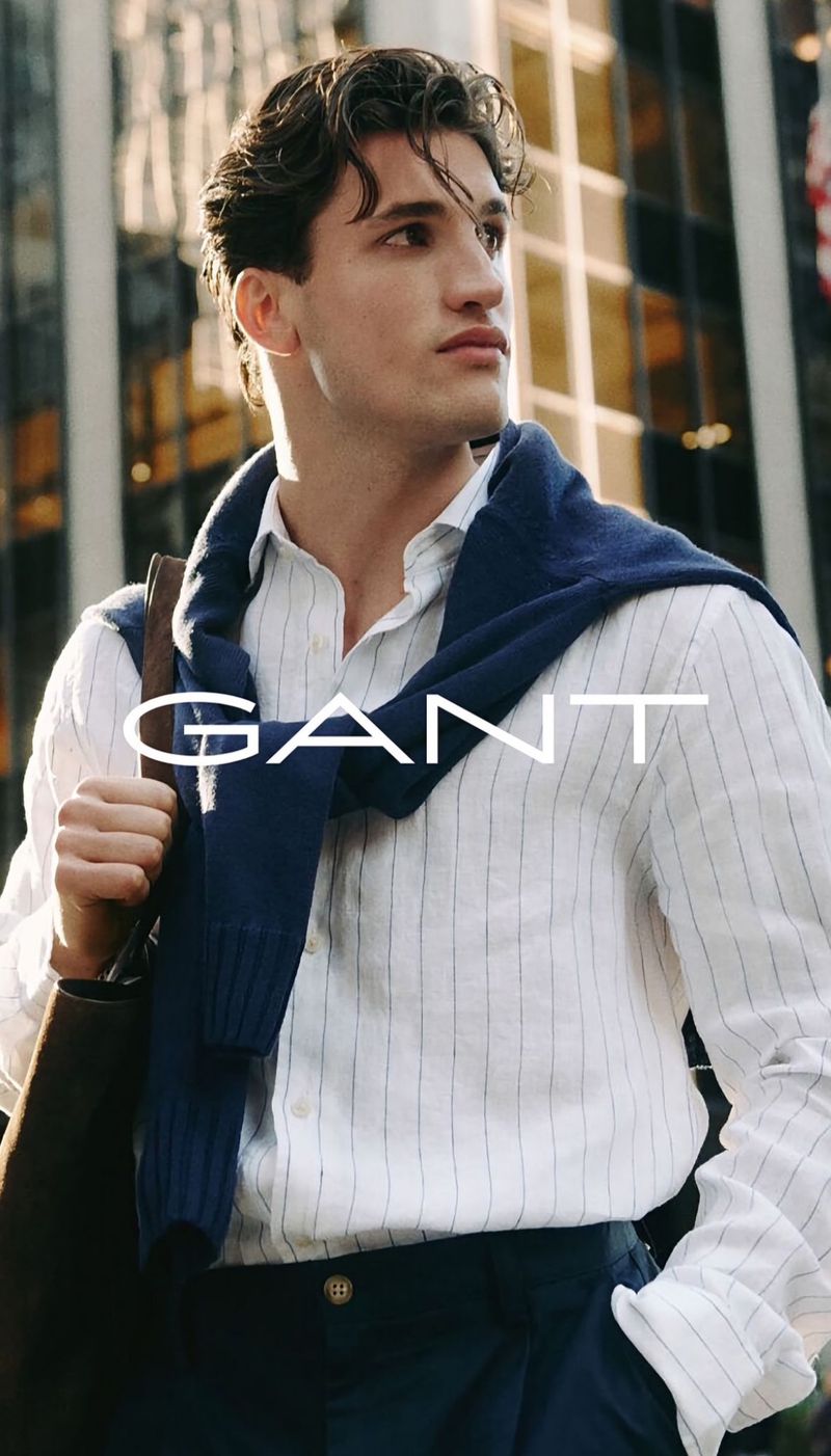 A young adult male wears a striped shirt and blue sweater with buildings in the background, highlighting fashion apparel and urban style.