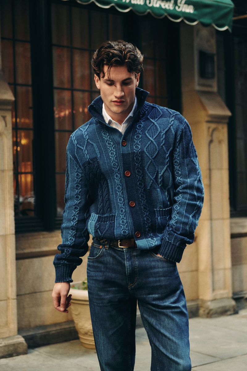 Man wearing a blue cable knit cardigan and jeans stands on a city street in front of an inn, showcasing casual men's fashion.