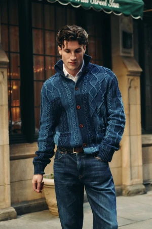 Man wearing a blue cable knit cardigan and jeans stands on a city street in front of an inn, showcasing casual men's fashion.