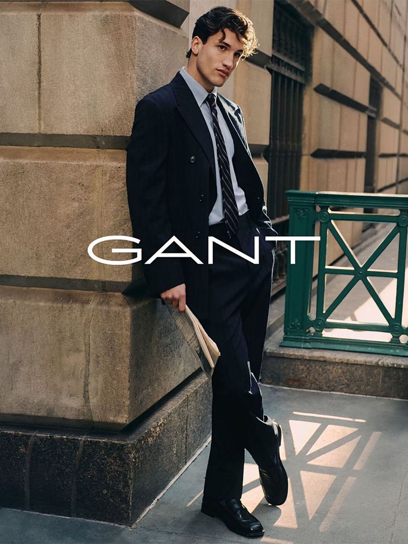 A stylish dark-haired man in a dark suit stands on a city street leaning against a stone wall in an advertisement for Gant clothing.