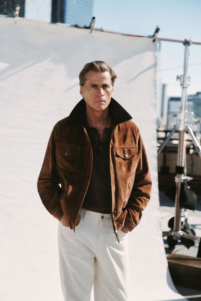 Portrait of a stylish adult man in a brown jacket and white pants, posing against a neutral backdrop with a blurred cityscape background.